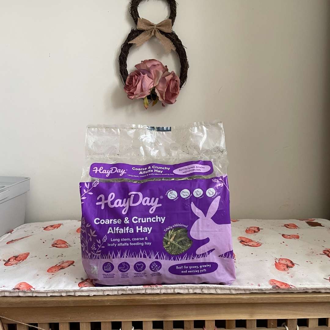 Purple package of HayDay Alfalfa Hay on a wooden surface with a decorative bunny wreath in the background.